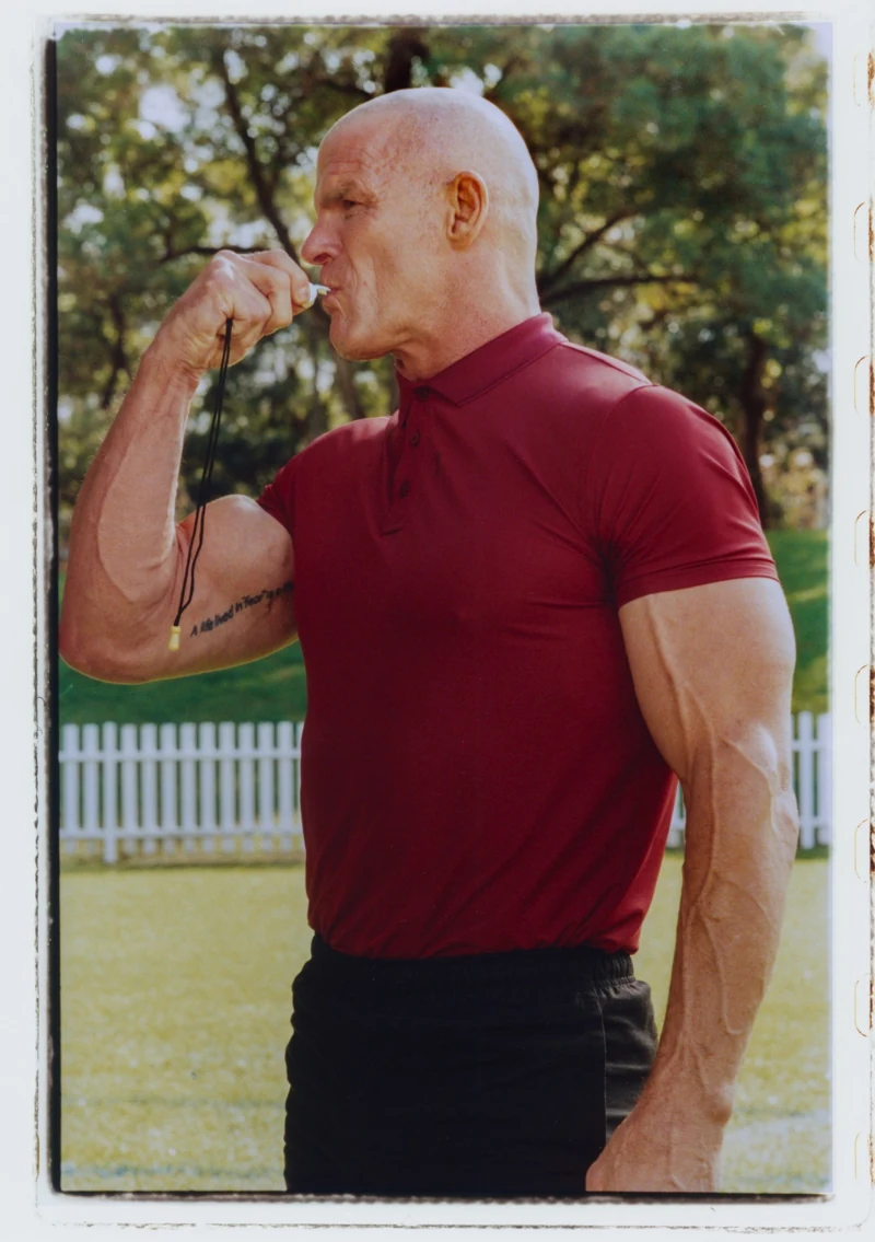 Ian Roberts in a red shirt on the sideline, holding a whistle to his mouth.