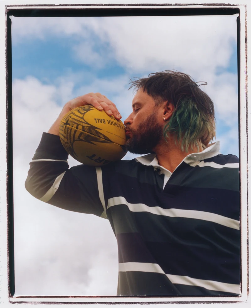 Mitch Brown kisses a yellow football, wearing a striped rugby shirt. Photography by Lula Cucchiara.